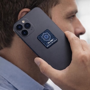 Puede incluir: Un smartphone gris oscuro con un escudo protector 5G EMF adherido a la parte trasera. El escudo es de forma cuadrada con un diseño azul y el texto "5G EMF PROTECTIVE SHIELD". El teléfono se sostiene cerca de la oreja.