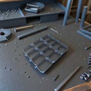 Op de afbeelding: Een grijze plastic organizer met 12 vakken voor kleine items. De organizer staat op een grijs oppervlak met kleine metalen stukjes die eromheen verspreid liggen.