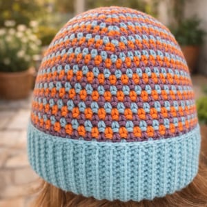 May include: A handmade beanie hat with a ribbed light blue brim and a patterned crown. The crown features a repeating design of orange, light blue, and purple squares. The hat is made of soft yarn.