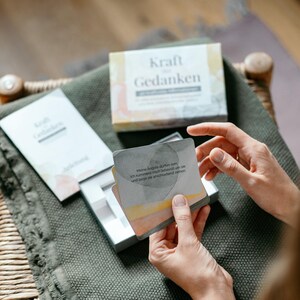 May include: A person holding a deck of cards with the words "Kraft Gedanken" on the box and cards. The cards have watercolor-style artwork and German text. The box and cards are on a green blanket, with a wooden chair in the background.