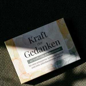 May include: A rectangular box with the German text "Kraft der Gedanken" and "40 kraftvolle Affirmationen" is displayed. The box has a watercolor design with orange and yellow hues. The box is on a textured green surface.