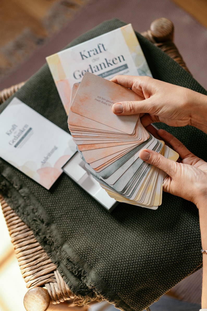 May include: A deck of cards with the German text "Kraft der Gedanken" and a book with the same title. The cards are held in a person's hands, fanned out to display the colorful designs. The cards have the text "Ich bin gesund mein Baby ist".