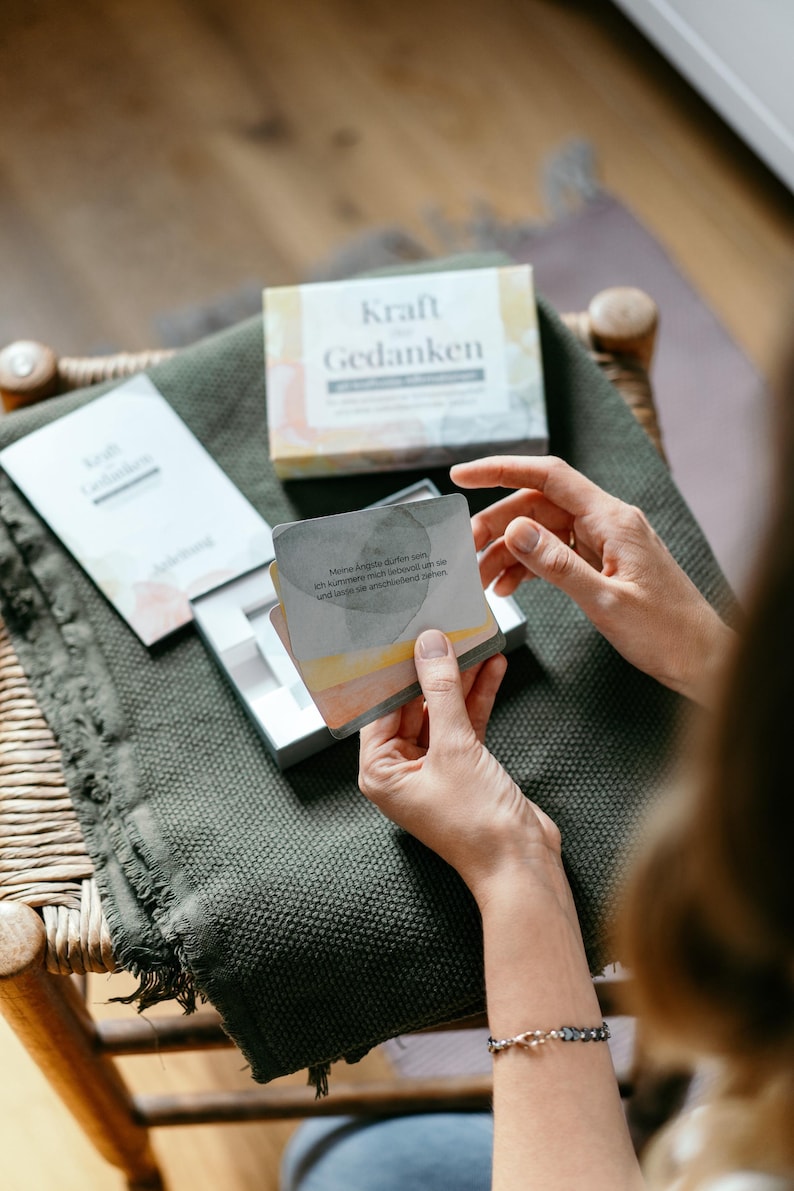 May include: A person holding a deck of cards with the words "Kraft Gedanken" on the box and cards. The cards have watercolor-style artwork and German text. The box and cards are on a green blanket, with a wooden chair in the background.