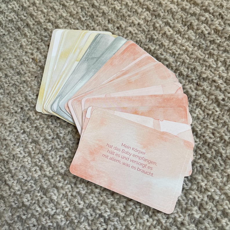 May include: A deck of cards with watercolor-style designs in shades of pink, yellow, and gray. The cards have rounded corners and feature text in German. The cards are fanned out on a textured surface.