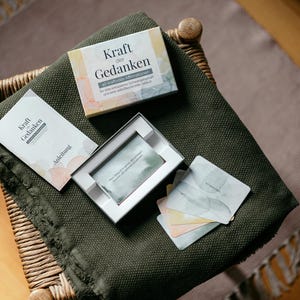 May include: A set of cards and a box with the German text "Kraft der Gedanken" (Power of Thoughts) on a green blanket. The box is white with watercolor-style pastel accents. Several cards are visible, along with a booklet.