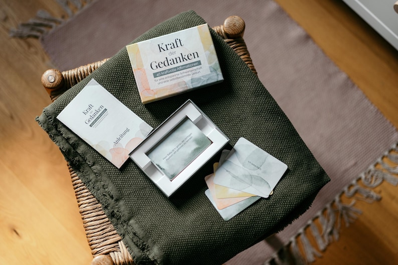 May include: A set of cards and a box with the German text "Kraft der Gedanken" (Power of Thoughts) on a green blanket. The box is white with watercolor-style pastel accents. Several cards are visible, along with a booklet.