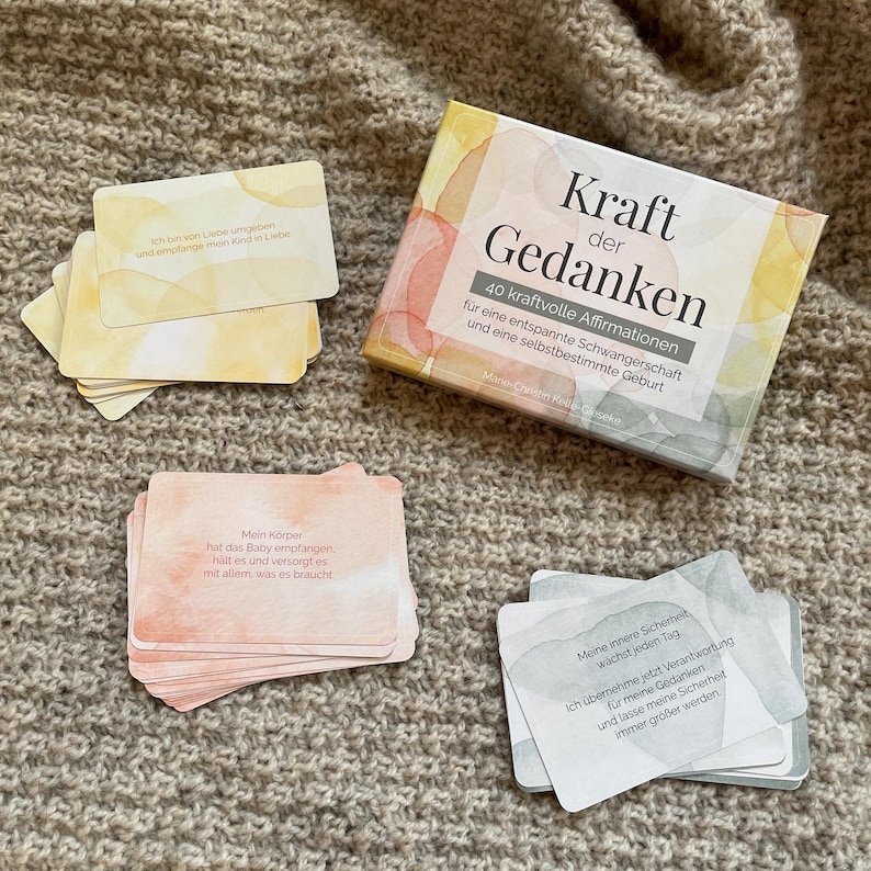 May include: A set of affirmation cards and a box titled "Kraft der Gedanken" with text in German. The cards have watercolor backgrounds in shades of yellow, peach, and gray. The box and cards are on a textured, neutral-toned surface.
