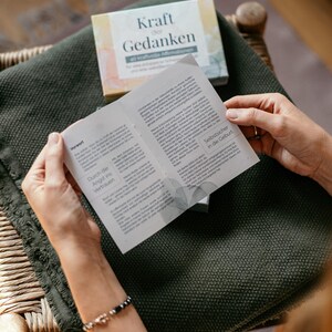 May include: A person is holding open a booklet with text, above a dark green blanket. A box with the words "Kraft der Gedanken" is visible. The booklet's pages are filled with text. A bracelet is visible on the person's wrist.