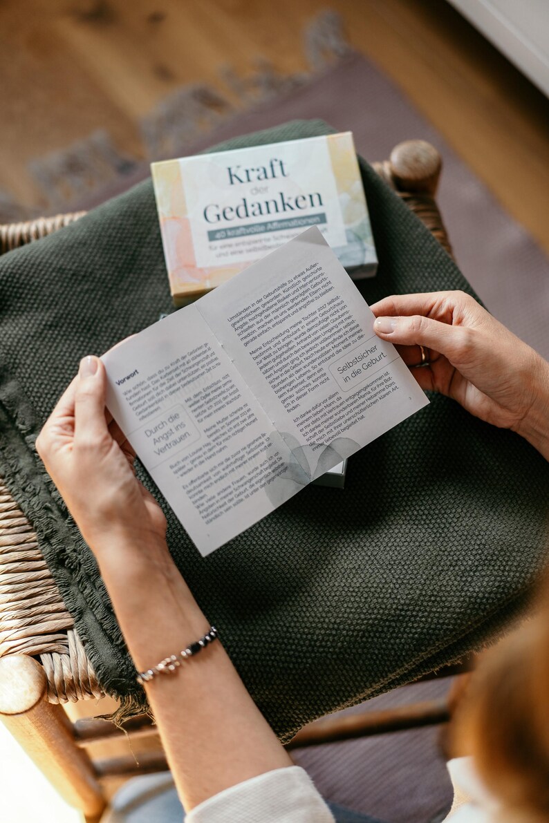 May include: A person is holding open a booklet with text, above a dark green blanket. A box with the words "Kraft der Gedanken" is visible. The booklet's pages are filled with text. A bracelet is visible on the person's wrist.