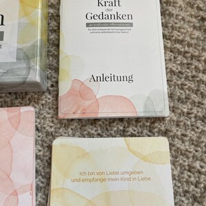 May include: A set of affirmation cards and a booklet with the title "Kraft der Gedanken" in German. The cards and booklet have watercolor-style designs in shades of yellow, orange, and pink. The text on the card reads "Ich bin von Liebe umgeben und empfange mein Kind in Liebe."