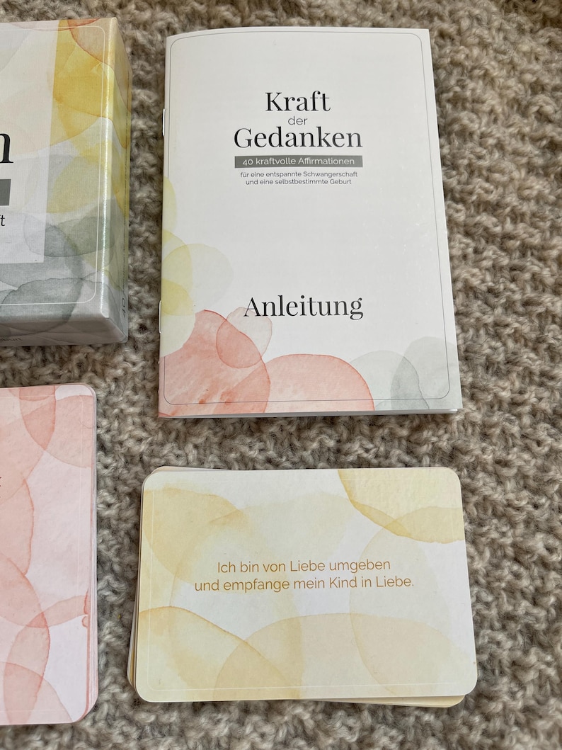 May include: A set of affirmation cards and a booklet with the title "Kraft der Gedanken" in German. The cards and booklet have watercolor-style designs in shades of yellow, orange, and pink. The text on the card reads "Ich bin von Liebe umgeben und empfange mein Kind in Liebe."