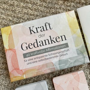 May include: A deck of affirmation cards titled "Kraft der Gedanken" (Power of Thoughts) with watercolor-style artwork. The box includes text in German, promoting a relaxed pregnancy and self-determined birth. Additional cards are visible.