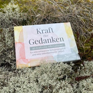 May include: A book titled "Kraft der Gedanken" (Power of Thoughts) with watercolor-style cover art. The book's title and affirmations are in German, with the subtitle mentioning a relaxed pregnancy and self-determined birth. The book is set on a bed of moss.