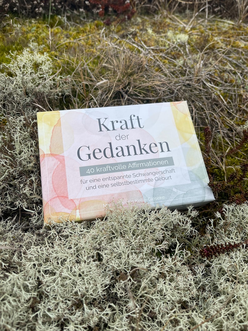 May include: A book titled "Kraft der Gedanken" (Power of Thoughts) with watercolor-style cover art. The book's title and affirmations are in German, with the subtitle mentioning a relaxed pregnancy and self-determined birth. The book is set on a bed of moss.