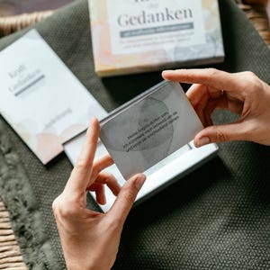 May include: A person holds a card from a set of "Kraft Gedanken" cards, with text in German. The card is gray with text, and the box is visible in the background. The box is white with the words "Kraft Gedanken" on it.