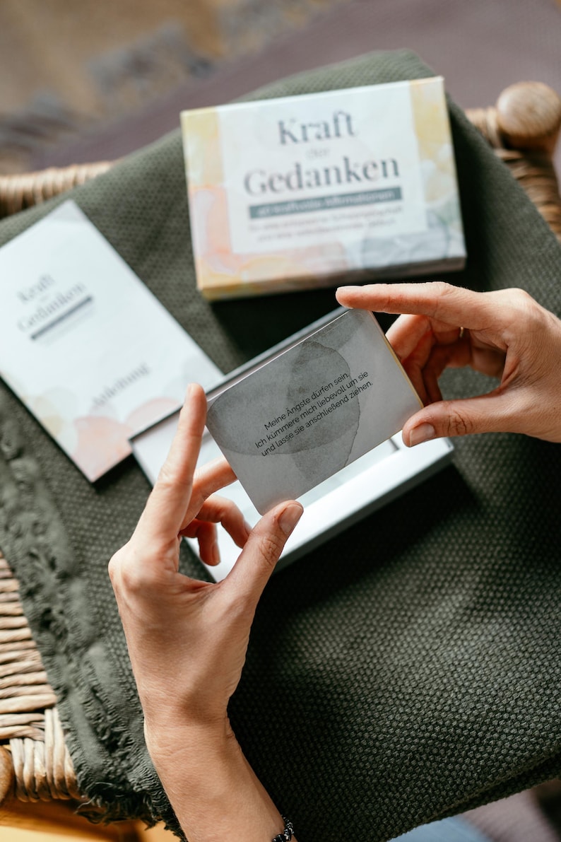 May include: A person holds a card from a set of "Kraft Gedanken" cards, with text in German. The card is gray with text, and the box is visible in the background. The box is white with the words "Kraft Gedanken" on it.