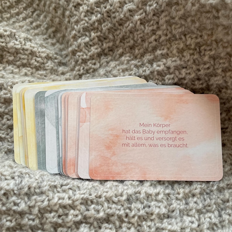 May include: A stack of pastel-colored cards with rounded corners, featuring watercolor-style backgrounds. The top card has text in German. The cards are resting on a textured, light brown surface.