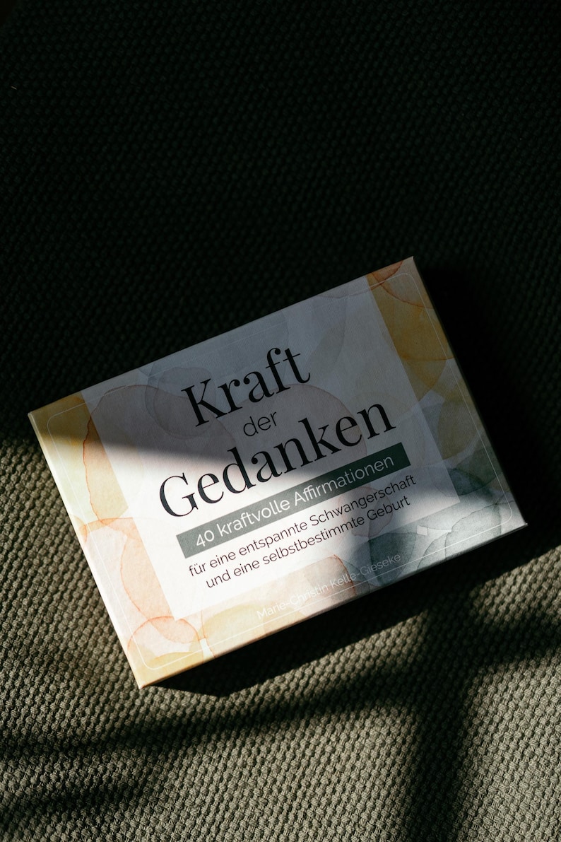 May include: A rectangular box with the German text "Kraft der Gedanken" and "40 kraftvolle Affirmationen" is displayed. The box has a watercolor design with orange and yellow hues. The box is on a textured green surface.