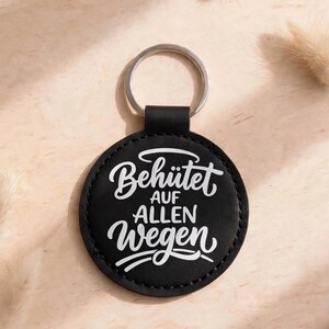 Könnte beinhalten: Ein runder schwarzer Schlüsselanhänger mit dem weißen Text Behütet AUF ALLEN Wegen. Der kreisförmige Anhänger besteht aus einem glatten Material mit sichtbaren Nähten am Rand und ist an einem silbernen Metallring befestigt.