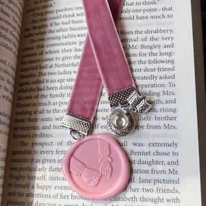 Könnte beinhalten: Ein staubrosa Samt-Lesezeichen mit silberfarbenen Metallakzenten und einem rosa Wachssiegel. Das Lesezeichen liegt auf einem offenen Buch. Das Wachssiegel zeigt zwei Hände, die sich umfassen.