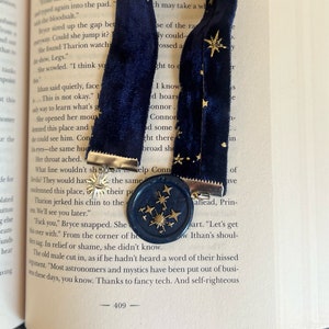 Könnte beinhalten: Marineblauer Samt-Lesezeichen mit goldenen Sternakzenten und einem passenden Wachssiegel. Das Lesezeichen liegt auf einem offenen Buch und verleiht dem Lesen eine elegante Note.