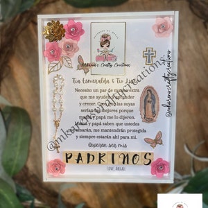 May include: A white frame with a pink and gold floral design. The frame contains a handwritten note with the text "Tia Esmeralda & Tio Luna" and "Quieren ser mis PADRINOS". The note is surrounded by pink flowers, a gold cross, and a rosary.