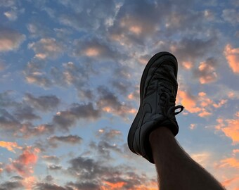 Könnte beinhalten: Ein Bein ist nach oben gestreckt und zeigt auf einen farbenfrohen Sonnenuntergangshimmel. Die Person trägt einen weißen Sneaker mit schwarzer Sohle.