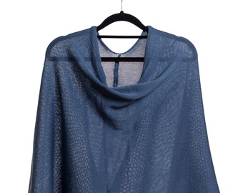 Puede incluir: Un poncho de punto azul colgado en una percha negra. La prenda presenta una textura fina y semitransparente con un cuello drapeado y un dobladillo asimétrico que cae en dos puntas distintas en la parte inferior de la tela.
