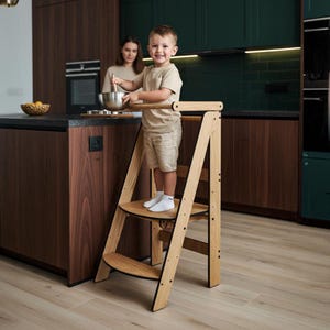 Könnte beinhalten: Ein hölzerner Küchen-Trittschemel mit einem Kind, das darauf steht. Der Trittschemel ist aus hellem Holz mit schwarzen Akzenten gefertigt. Das Kind lächelt und hält eine Schüssel und einen Löffel. Im Hintergrund ist eine Frau zu sehen.