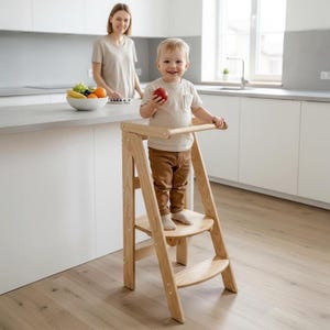 Peut inclure: Un tabouret en bois naturel pour enfant avec une barre de sécurité. Le tabouret a deux marches et une base incurvée. Un jeune enfant se tient sur la marche supérieure, tenant une pomme. Une femme est en arrière-plan.