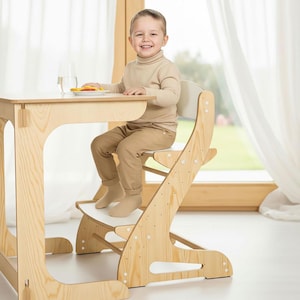 Op de afbeelding: Een lichte houten kinder-tafel en stoel set. De stoel heeft een witte zitting en voetensteun. De tafel heeft een lichte houten afwerking. Een jong kind zit lachend aan de tafel. De tafel en stoel staan bij een raam met witte gordijnen.