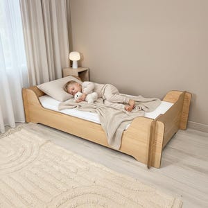 Puede incluir: Una cama infantil de madera clara con paneles laterales curvos y un marco de perfil bajo. La cama está vestida con un colchón blanco, una almohada beige y una manta de punto beige a juego. La estructura presenta un acabado de veta natural.
