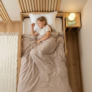 Puede incluir: Una estructura de cama de madera clara con listones verticales, que incluye una almohada blanca y un edredón beige liso. Una persona duerme en la cama con una camiseta blanca. Una pequeña mesita de noche de madera con una lámpara está al lado.