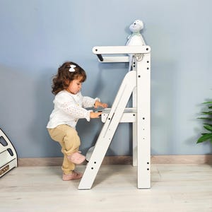 Puede incluir: Un taburete de madera blanco con una pequeña plataforma y un diseño de escalera. Un niño sube los escalones. Un peluche está sentado en la plataforma. El taburete está diseñado para niños.