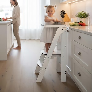 Pode incluir: Um banco de cozinha de madeira branca com dois degraus e uma barra de segurança. Uma criança pequena com um vestido branco está em pé no banco, sorrindo e segurando um utensílio. Uma mulher é vista ao fundo, preparando comida na bancada da cozinha.