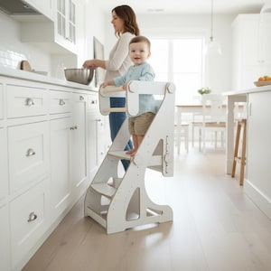 Può includere: Una torre di apprendimento in legno bianco con piattaforma regolabile su più livelli e ringhiera di sicurezza. La struttura presenta una base robusta con pannelli laterali, un gradino per salire e una barra di supporto superiore.
