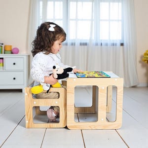 Peut inclure: Un ensemble table et chaise pour enfants en bois clair. La chaise est intégrée à la table. Un enfant est assis sur la chaise, tenant une peluche Mickey Mouse et regardant un puzzle coloré sur la table.