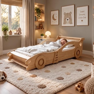 Op de afbeelding: Een lichtgekleurd houten bed in de vorm van een auto met een wit matras en beddengoed. Het frame heeft uitgesneden wielontwerpen en een lage structuur. Een persoon slaapt op het bed, dat op een crèmekleurig kleed met bruine stippen staat.