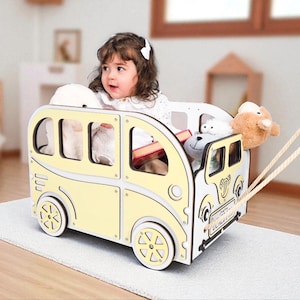 Pode incluir: Um autocarro de brinquedo de madeira amarelo e branco, com uma criança dentro. O autocarro tem bordas arredondadas, janelas e rodas. Está cheio de bichos de pelúcia e tem uma corda para puxar. O autocarro está sobre um tapete claro.