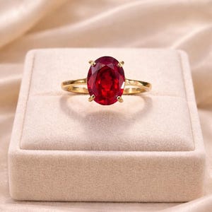 Può includere: Un anello d'oro con una grande pietra preziosa rosso scuro a taglio ovale. L'anello è incastonato in una classica montatura a quattro griffe e poggia su un portagioie in velluto beige su uno sfondo di tessuto satinato.
