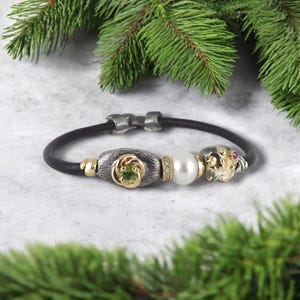 Könnte beinhalten: Ein schwarzes Lederarmband mit silbernen und goldenen Akzenten. Das Armband hat eine silberne Perle mit einem grünen Stein, eine Perlenperle und eine goldene Perle mit einem roten Stein. Der Verschluss ist silberfarben.