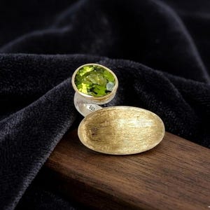 May include: A sterling silver ring with a gold-plated band and a large, oval, light green gemstone. The ring has a textured gold-plated accent. The ring is displayed on a dark wooden surface.