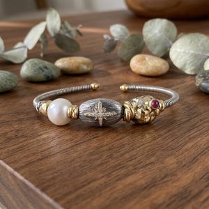 Peut inclure: Un bracelet manchette composé d'une bande argentée torsadée ornée d'une perle blanche, d'une perle ovale texturée foncée avec une étoile dorée incrustée de petites pierres claires, et d'une perle dorée irrégulière avec une petite pierre rouge.