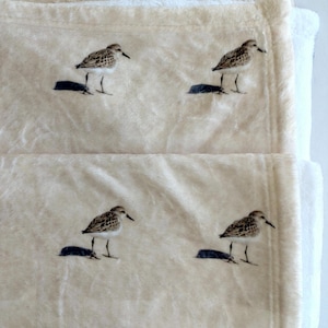 May include: Cream-colored plush blanket featuring four embroidered shorebird designs. Each bird is depicted with brown and white patterned feathers, a black beak, and dark blue shadows. The blanket has a soft, textured appearance.