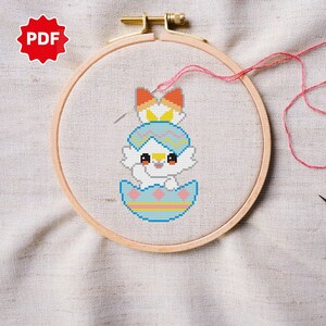 Peut inclure: Modèle de point de croix d'un lapin blanc portant un chapeau d'œuf de Pâques bleu et jaune, assis dans un œuf de Pâques bleu et jaune. Le motif est dans un cerceau de broderie en bois.