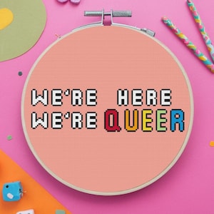 May include: A pink embroidery hoop with the phrase "We're here, We're queer" in rainbow pixel art.