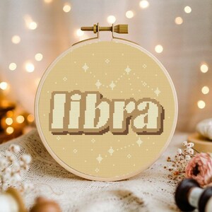 May include: A beige embroidery hoop with a cross-stitch design of the word "libra" in a retro font with a constellation of stars in the background.
