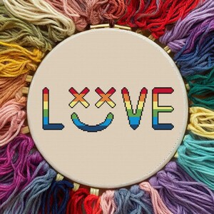 Peut inclure: Un cercle de broderie au point de croix avec le mot "LOVE" en couleurs arc-en-ciel. Les lettres sont brodées sur un tissu beige, entourées de fil à broder coloré dans diverses nuances de rouge, orange, jaune, vert, bleu et violet.