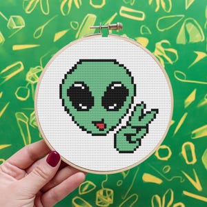 Peut inclure: Motif de point de croix représentant un extraterrestre vert faisant un signe de paix. L'extraterrestre a de grands yeux et la langue qui sort.