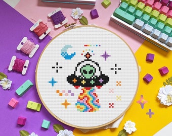 Op de afbeelding: Een kleurrijk kruissteekborduurwerk met een alien in een UFO met een regenboogstraal. Het ontwerp bevat sterren, een maan en een regenboog. De borduursel zit in een houten hoepel, omringd door naaibenodigdheden en toetsenbordtoetsen.